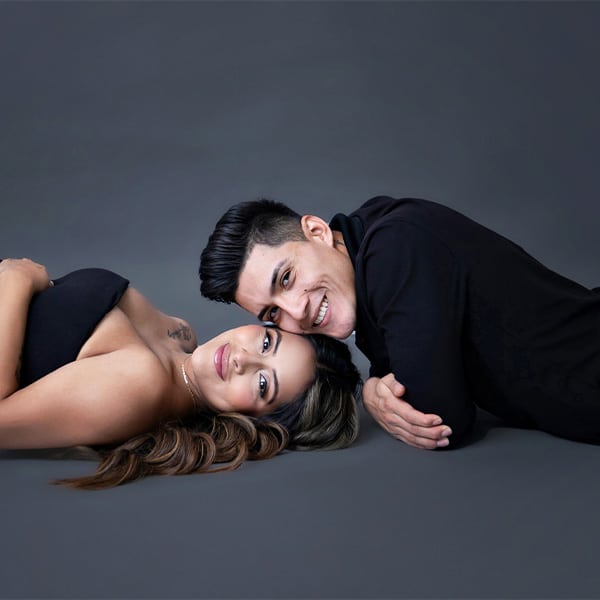 A pregnant couple lying together on the floor, smiling warmly at the camera, with the woman wearing a black dress and the man dressed in black against a gray background.