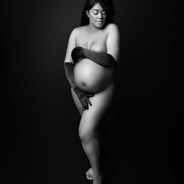 A black and white portrait of a pregnant woman wearing gloves and a decorative headpiece, posing against a dark background.