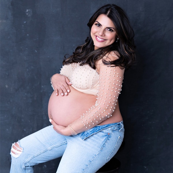 A smiling pregnant woman sitting on a stool, wearing ripped jeans and a pearl-embellished top, cradling her belly against a textured dark background.