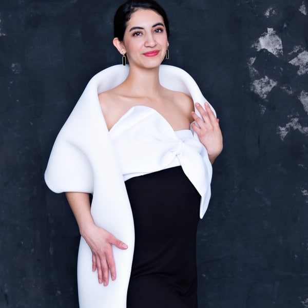 A pregnant woman wearing a modern white wrap and black dress, posing gracefully against a textured dark background.