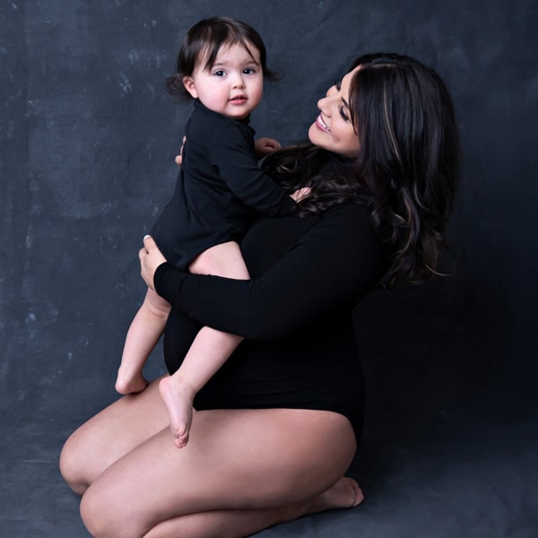 A pregnant woman in a black bodysuit kneeling on the floor, smiling while holding her toddler against a dark background.