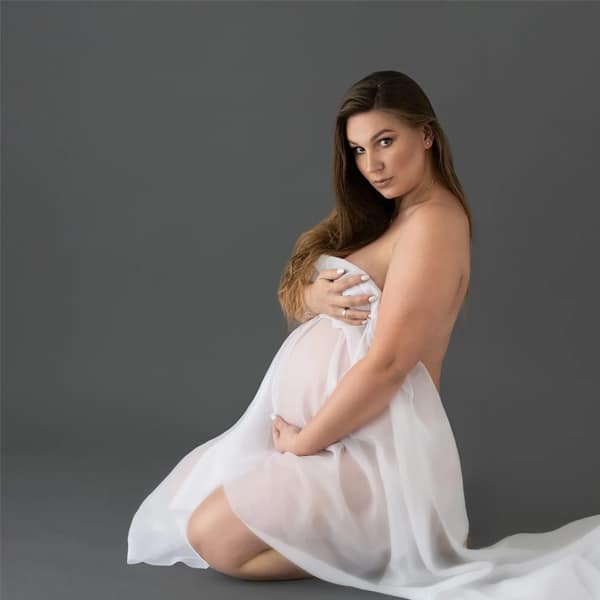 A pregnant woman kneeling on the floor, draped in a flowing white gown, holding her belly and gazing toward the camera against a gray background.