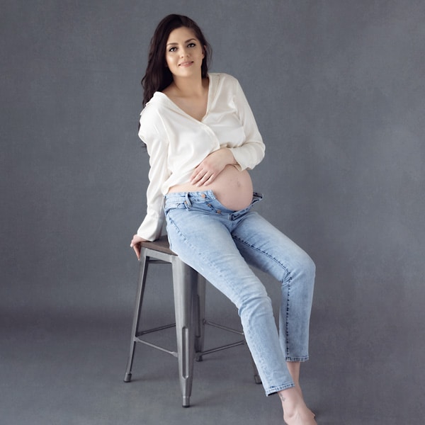 A pregnant woman sitting on a metal stool, wearing a white blouse and jeans, gently holding her belly against a gray studio background.