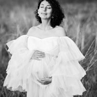 A pregnant woman in a flowing off-the-shoulder tulle gown, standing outdoors in a grassy field with her hands gently cradling her belly and her eyes closed.