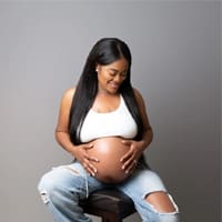 A pregnant woman sitting on a wooden stool, wearing ripped jeans and a white tank top, smiling as she cradles her belly against a gray background.