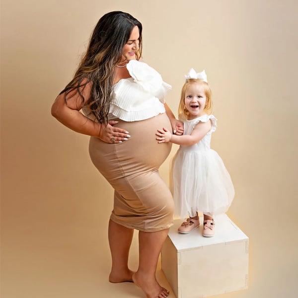 A pregnant woman in a fitted beige dress standing beside her young daughter dressed in white, smiling and holding her mother's belly in a studio with a soft beige background.