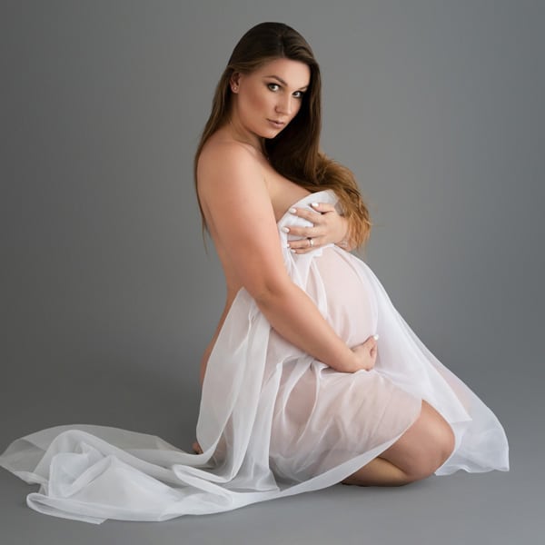 A pregnant woman kneeling on the floor, wrapped in soft white fabric, posing against a gray background.