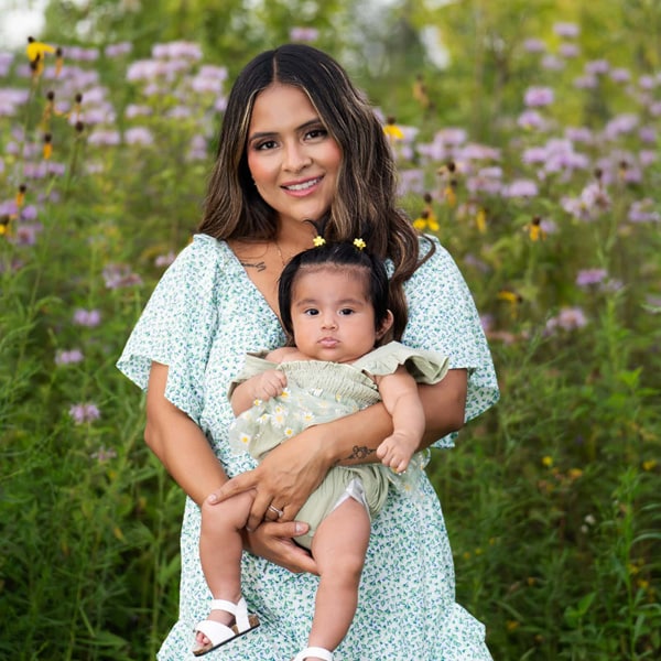 A smiling mother in a floral dress holding her baby in a wildflower field with purple and yellow blossoms.