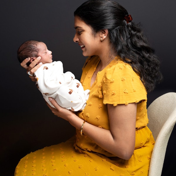 A smiling mother in a yellow dress lovingly holding her newborn baby wrapped in a white blanket, against a dark background.