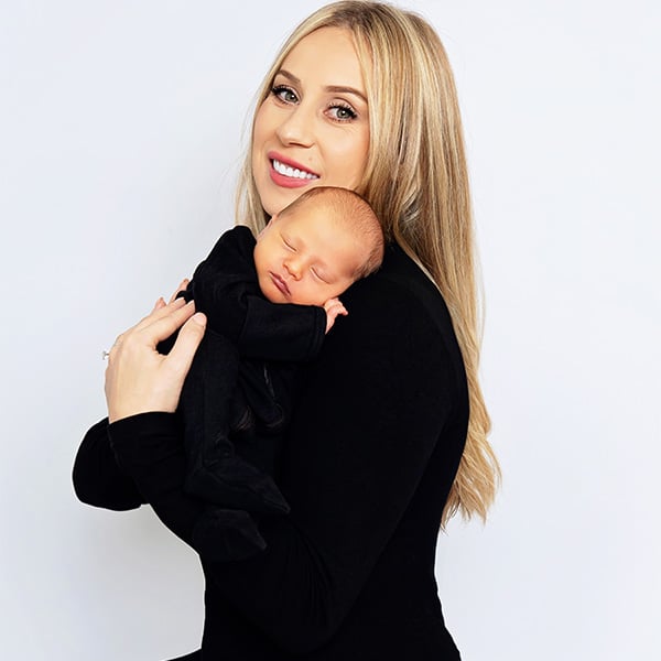 A smiling mother with long blonde hair holding her sleeping newborn baby dressed in black, against a light background.