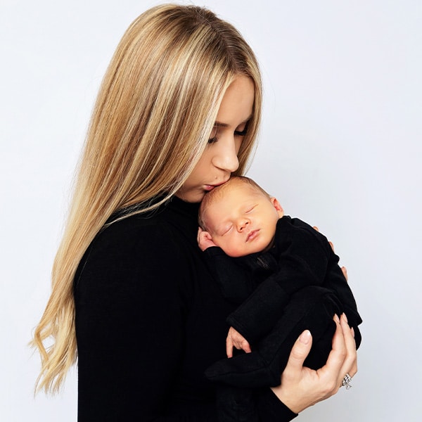 A mother with long blonde hair gently holding and kissing her sleeping newborn baby, both dressed in black, against a white background.