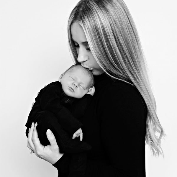 A mother gently kissing her newborn baby while holding them close in a black-and-white portrait.