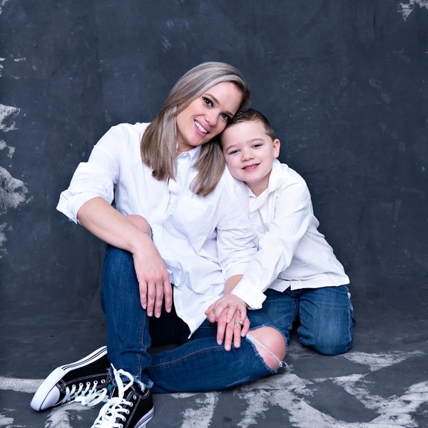 A mother and her young son sitting close together, dressed in white shirts and jeans, with a dark textured background.