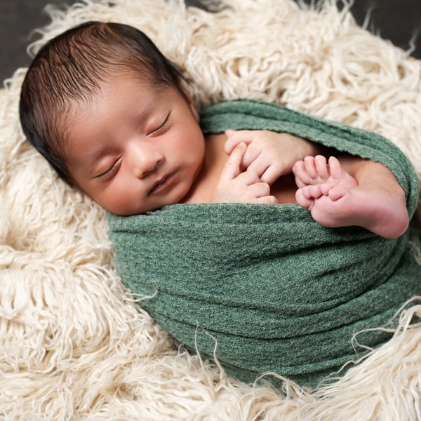 A sleeping newborn baby wrapped in a green cloth, lying on a fluffy cream-colored blanket.