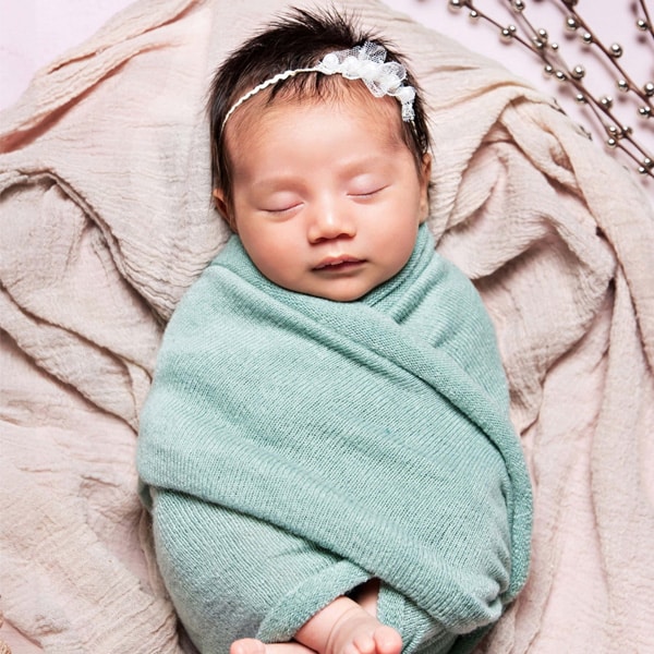 A sleeping newborn baby swaddled in a soft green cloth, lying on beige fabric with a white floral headband.