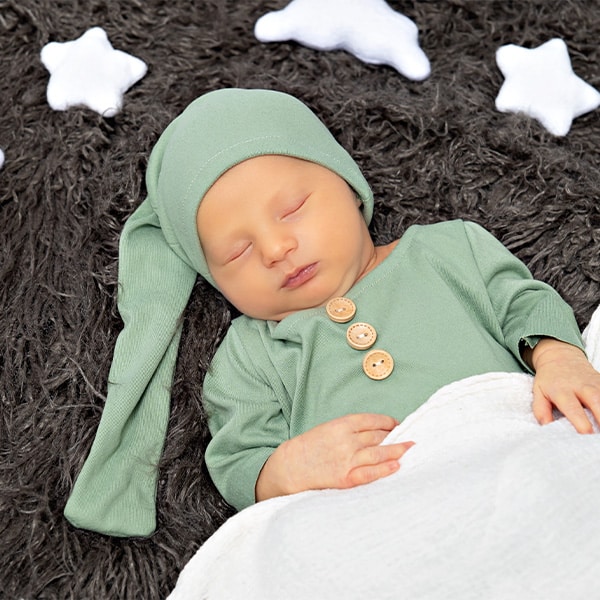 A smiling baby dressed as a little aviator, wearing suspenders and a pilot hat, lying on a green backdrop.