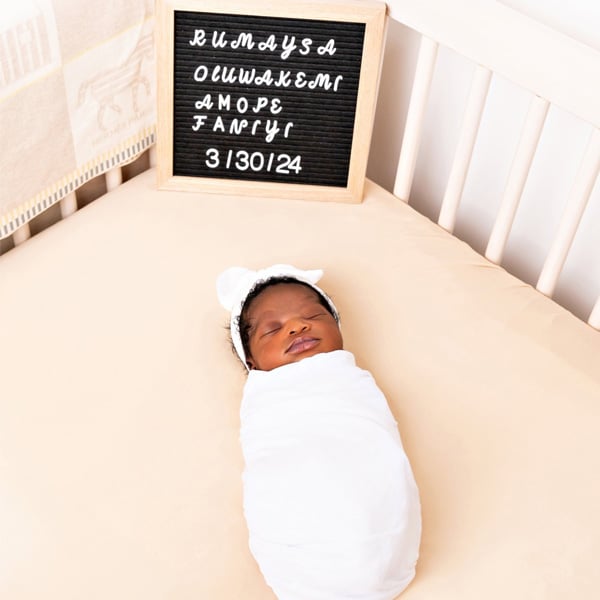 A newborn baby swaddled in white, sleeping in a crib with a name announcement board and date beside them.