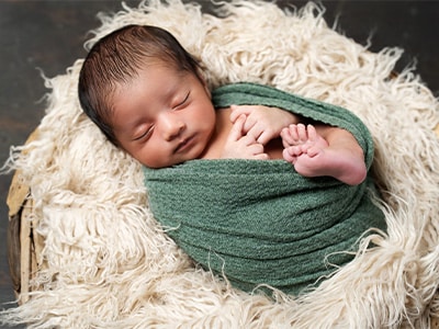 A peaceful newborn baby wrapped in a green blanket, sleeping in a wooden basket lined with soft cream-colored fur