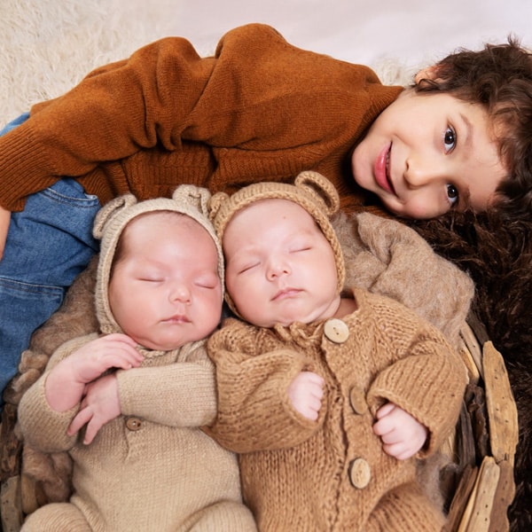 An older sibling smiling while lying next to newborn twins dressed in knitted bear-themed outfits.