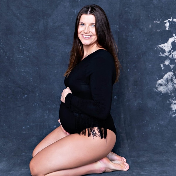 A pregnant woman kneeling on the floor, dressed in a black bodysuit with fringe details, smiling at the camera against a textured dark background.