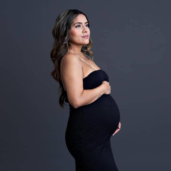 A pregnant woman in a black form-fitting dress, holding her belly while standing in profile against a gray background.