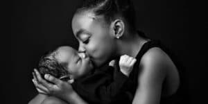 A young sister lovingly kissing her newborn sibling while sitting and cradling the baby.