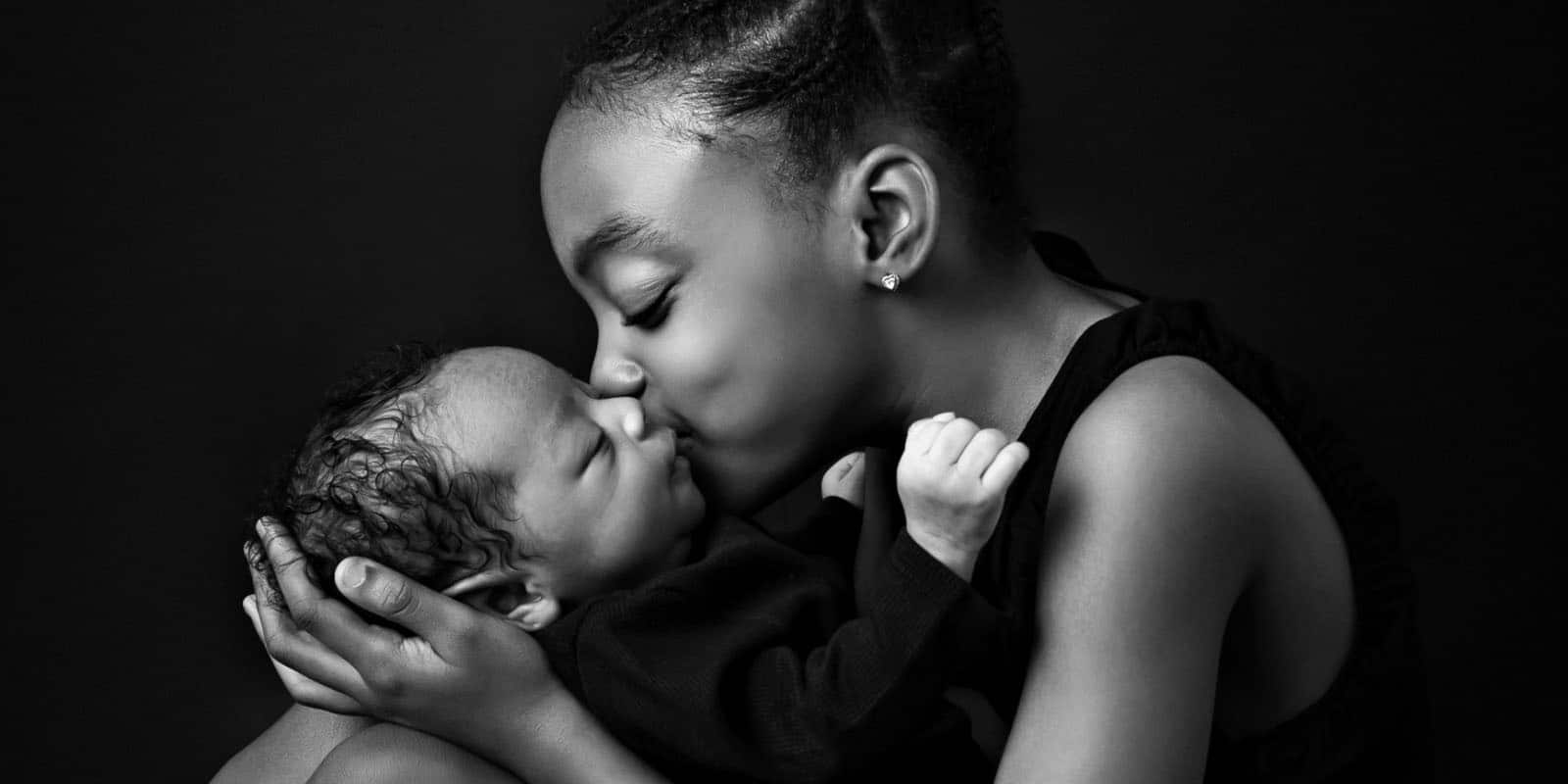 A young sister lovingly kissing her newborn sibling while sitting and cradling the baby.