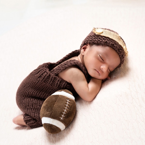 A newborn baby sleeping on a cream blanket, dressed in a brown knitted outfit and hat, with a small plush football beside them.