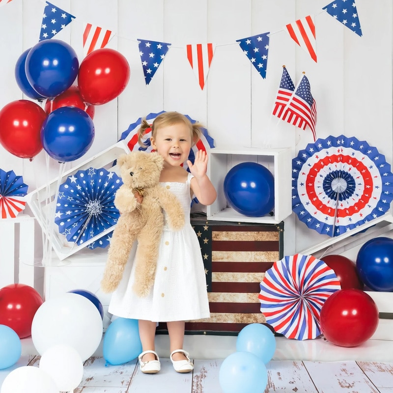 A toddler in a white dress holding a teddy bear, surrounded by red, white, and blue patriotic decorations and balloons for a Fourth of July celebration.