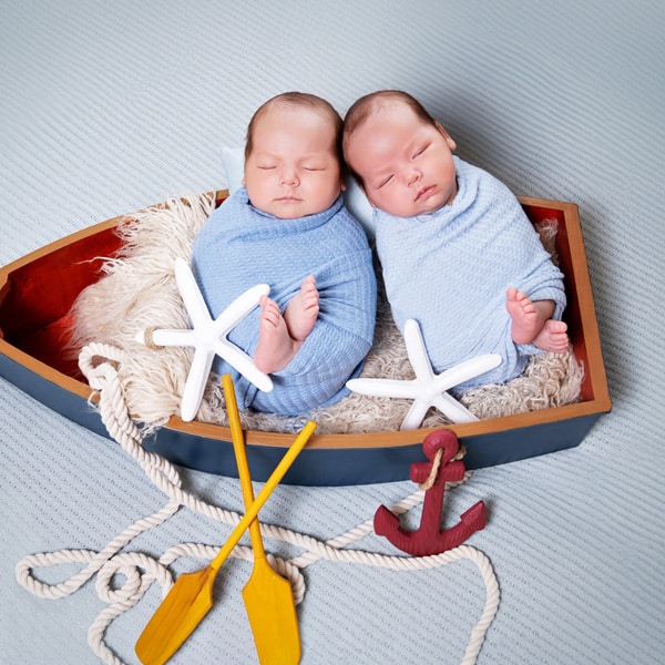 Two sleeping newborn twins wrapped in blue swaddles, lying inside a small wooden boat decorated with starfish and oars.