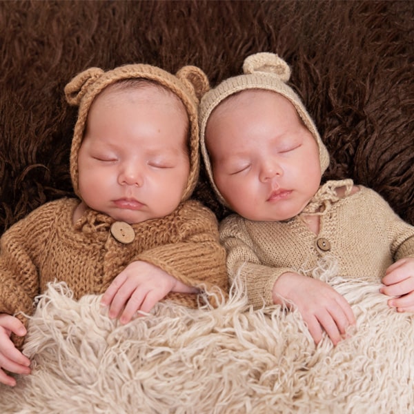 Two sleeping twin babies dressed in knitted bear-themed outfits, lying on a soft blanket.